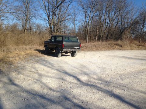 1979 Ford Bronco - A dark 1979 Ford Bronco is seen from the rear, parked on a gravel lot with trees in the background.