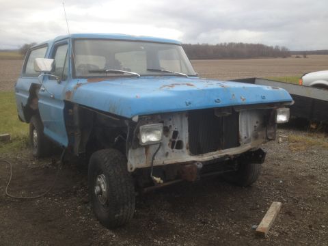 1979 Ford Bronco - A dilapidated blue Ford Bronco sits in a field, missing body panels and showing significant wear.