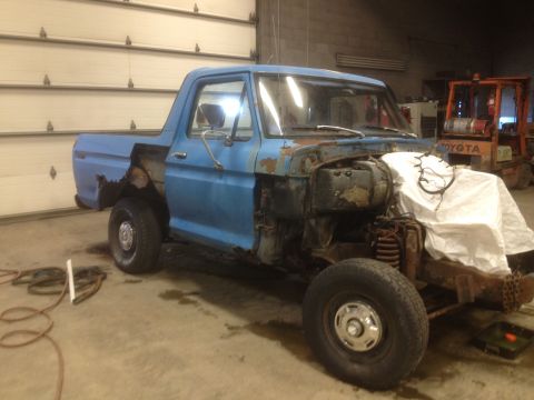 1979 Ford Bronco - This blue pickup truck is missing its front fenders and hood, revealing rusted engine components.