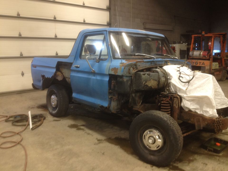 Blue pickup truck with front end disassembled, covered with a white sheet. A forklift is in the background