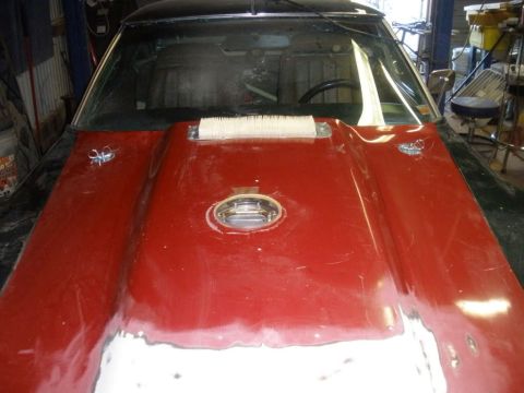 1976 Chevrolet Corvette - The front of a car, likely undergoing repairs, is shown with a prominent red hood and a central gas cap.