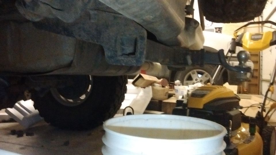 Underside rear view of a vehicle showing hitch ball, exhaust, and rear tire in a garage with a bucket and mower nearby