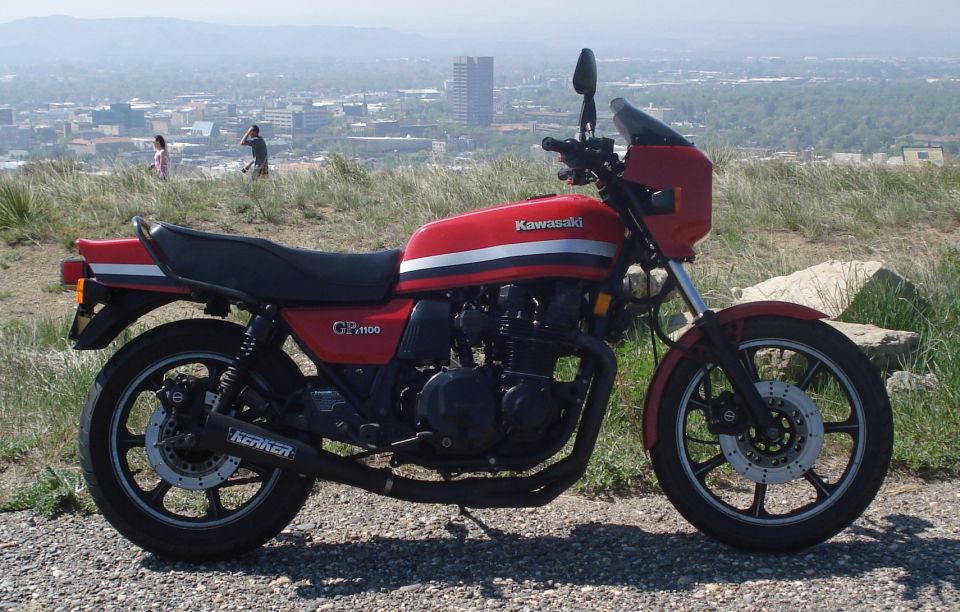 Red Kawasaki GP1100 motorcycle parked on gravel with a city skyline in the background