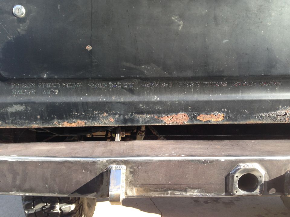 Close-up of a black vehicle panel with embossed text and rust spots. A metal bumper is visible below