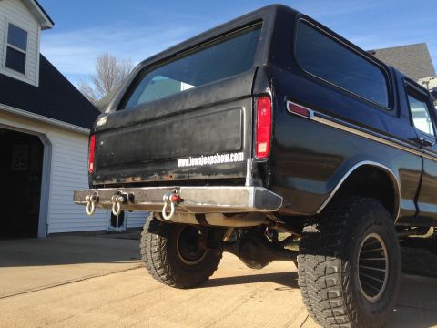 1979 Ford Bronco - The rear of a black Ford Bronco is shown with a custom bumper and large tow hooks. A website address is visible on the tailgate.