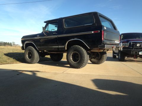 1979 Ford Bronco - A black SUV with large tires is parked on a concrete surface next to another vehicle.