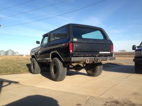 1979 Ford Bronco - Rear view of a lifted black Ford Bronco a custom bumper and tow hooks. A second vehicle is parked nearby.
