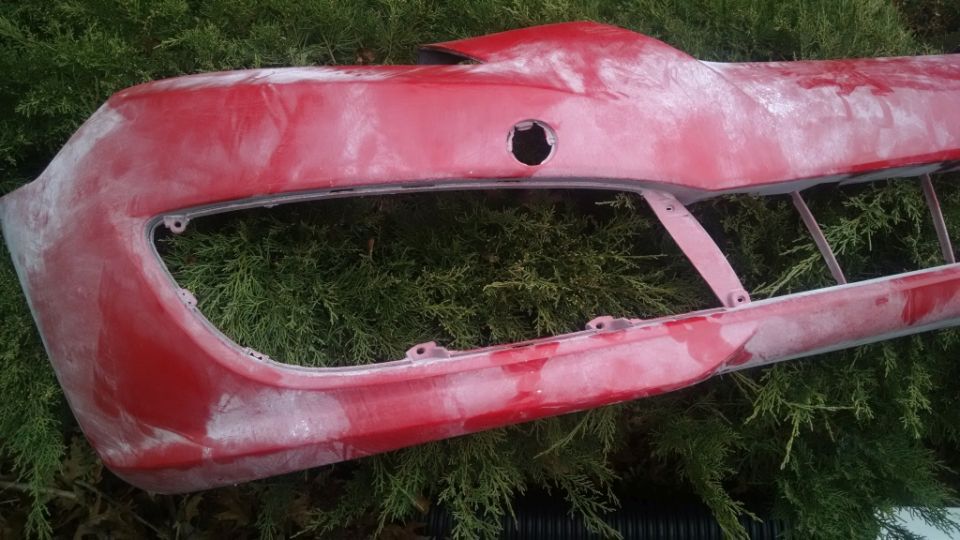 Red car bumper with white primer, resting on green foliage.