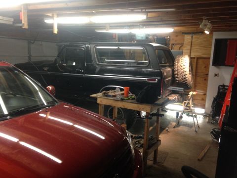 1979 Ford Bronco - A black Ford Bronco is parked in a garage next to a red car. A spare tire is mounted on the rear of the Bronco, and a workbench with tools…