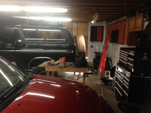 1979 Ford Bronco - A black Ford Bronco is parked in a garage next to a red vehicle. A workbench with tools and a spare tire is set up in front of the Bronco.