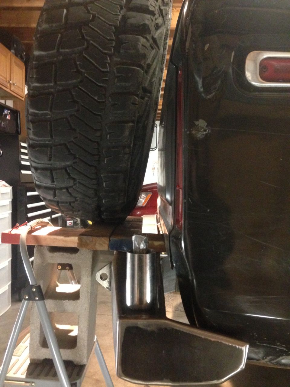 Rear corner of a dark SUV with a spare tire mounted, supported by cinder blocks and a custom bumper.