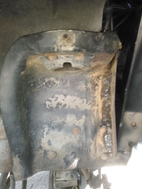1996 Ford Bronco - This image shows a heavily rusted and corroded metal bracket, likely part of a vehicle's undercarriage. The bracket is black with significa…