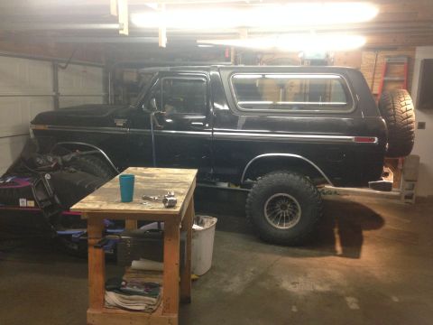 1979 Ford Bronco - A black 1970s Ford Bronco is parked in a garage next to a workbench. The vehicle has a spare tire mounted on the rear and large off-road ti…