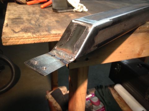 1979 Ford Bronco - A close-up shot shows a metal bracket with visible welds and drilled holes, resting on a wooden workbench. Various tools and items are blur…