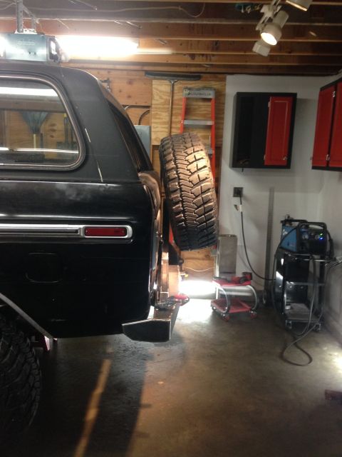 1979 Ford Bronco - The rear driver's side of a black SUV is shown in a garage setting, with a spare tire mounted to the back. A welding machine and other tool…