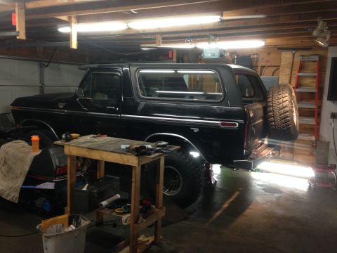 1979 Ford Bronco - A black 1979 Ford Bronco is parked in a garage with a spare tire mounted on the rear. A workbench with tools sits in front of the vehicle.