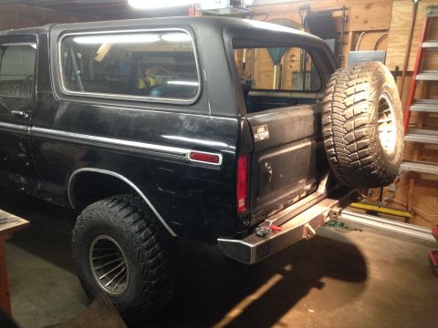 1979 Ford Bronco - The rear passenger side of a black Ford Bronco is shown in a garage. A spare tire is mounted on the rear bumper, and the vehicle has large…