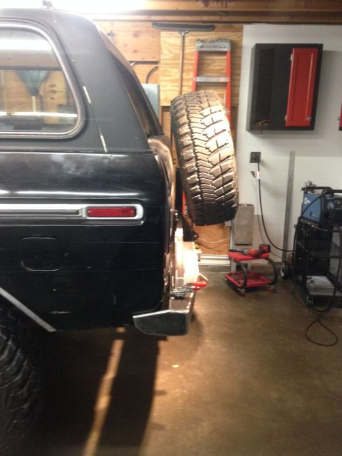 1979 Ford Bronco - The rear passenger side of a black SUV is shown mounted with a spare tire on a swing-away carrier. The vehicle is in a garage with tools an…