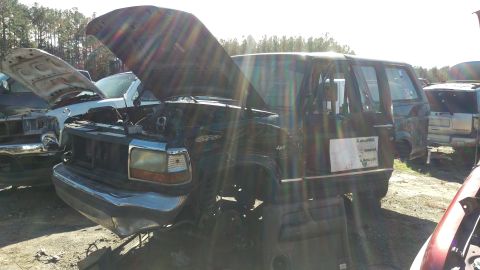 1994 Ford Bronco - A dark-colored, four-door SUV with its hood open is parked in a junkyard. Another vehicle with its hood open is visible to the left, and a…