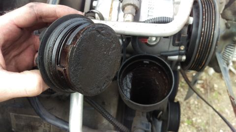 1994 Ford Bronco - A hand holds a very dirty oil filter cap next to the open housing of the oil filter, revealing the dirty interior. The engine bay of a 1994…