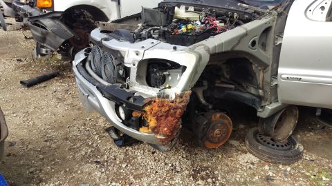 1994 Ford Bronco - The front end of a heavily damaged silver vehicle, likely a SUV, is shown in a junkyard setting. The front bumper is severely rusted and br…