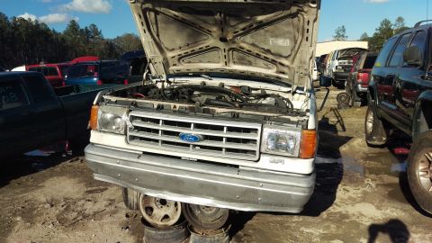 1994 Ford Bronco - The front end of a white Ford pickup truck is shown with its hood open, revealing the engine bay. The truck is parked in a junkyard with ot…