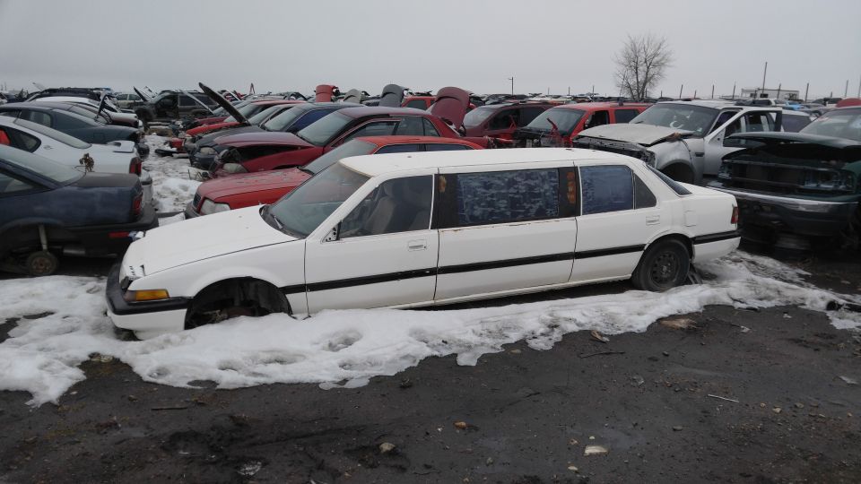 White stretch limousine in a junkyard, missing front wheels and hood.