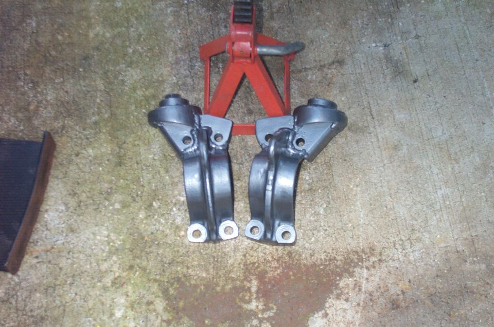 Two gray suspension brackets with bolts and holes, next to a red jack stand on concrete.