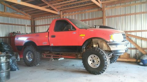 1982 Ford Bronco - A modified red pickup truck with oversized tires and a lifted suspension is parked inside a wooden barn.