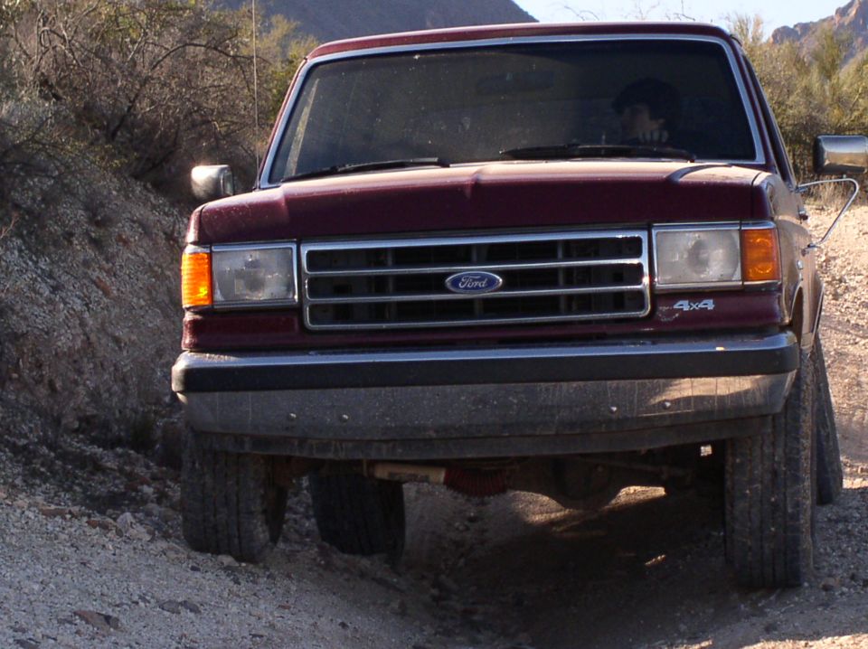 Front view of a maroon Ford Bronco driving on a dirt road. It has a chrome grille and 4x4 badging