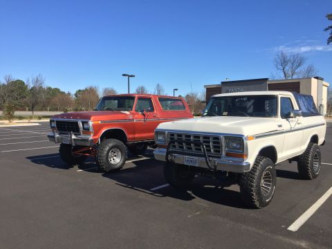 1979 Ford Bronco - Two lifted Ford Broncos are parked side-by-side in a parking lot on a clear day.