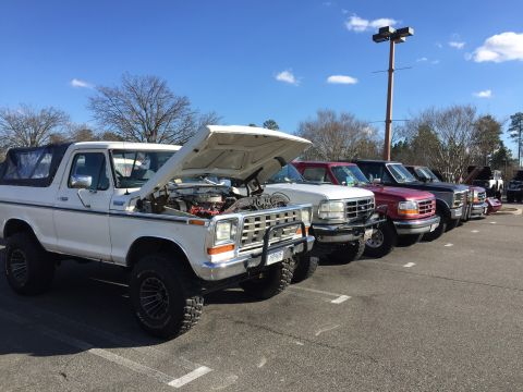 1979 Ford Bronco - A line of trucks and SUVs are parked in a lot under a bright blue sky, with the first vehicle being a white 1979 Ford Bronco with its hood…