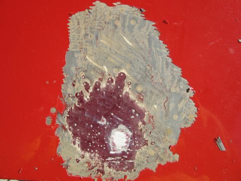 1976 Chevrolet Corvette - Close-up of damaged red car paint, exposing primer and metal, with visible texture from the impact.