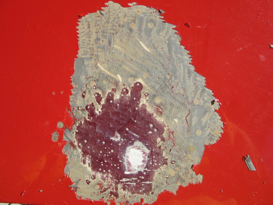 Red car panel with chipped paint revealing primer and underlying material, showing signs of impact