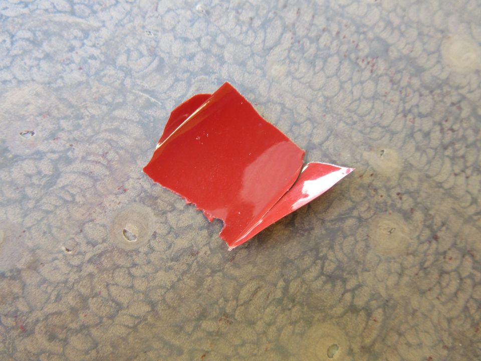 Red paint chip on a textured, light-colored surface. A white backing is visible on one edge
