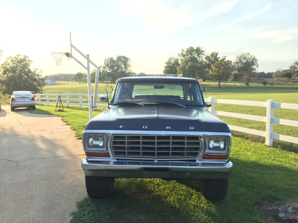 Front view of a dark blue Ford truck parked in a driveway with a basketball hoop behind it. Another car is visible in the background