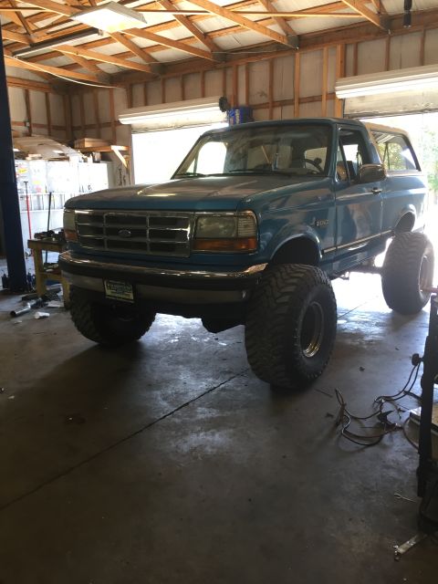 1995 Ford Bronco - A lifted, teal 1995 Ford Bronco with large off-road tires is parked inside a garage. The vehicle is viewed from a front 3/4 angle, showing…