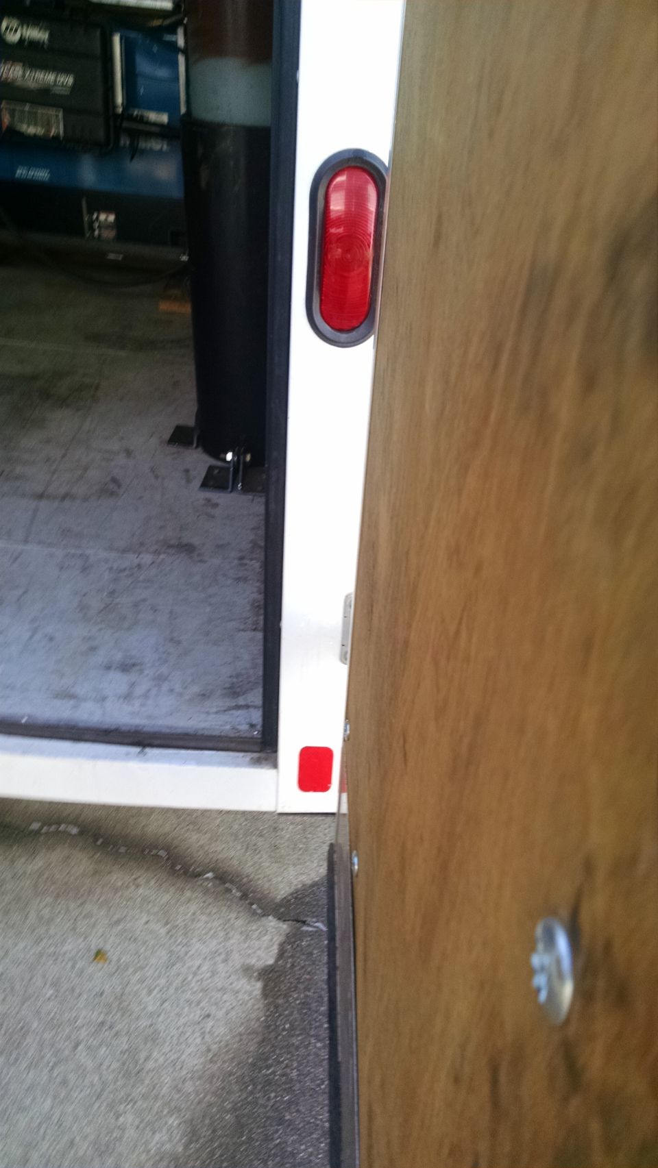 Rear corner of a white trailer with red oval light and reflector, wood door partially open.