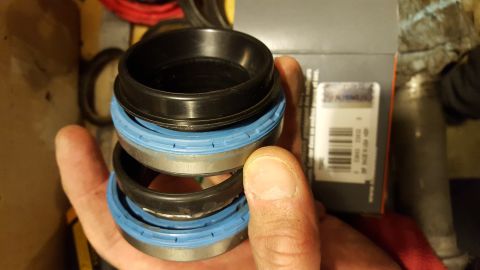 1994 Ford Bronco - A hand holds a stack of three black and blue fork seals, likely for a vehicle suspension system. A partial view of a product box with a bar…