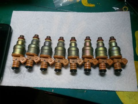 1996 Ford Bronco - A set of eight fuel injectors are laid out in a row on a white paper towel. The injectors are mostly metallic with orange plastic connector…