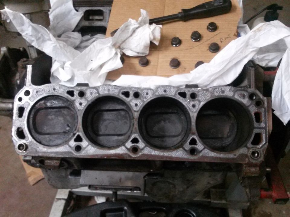 V8 engine block with four cylinder bores, bolts, and screwdriver.
