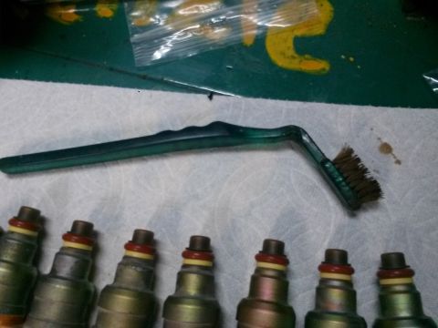 1996 Ford Bronco - A green-handled brush with brown bristles is positioned above a row of fuel injectors. The injectors are metallic with red and yellow O-rin…