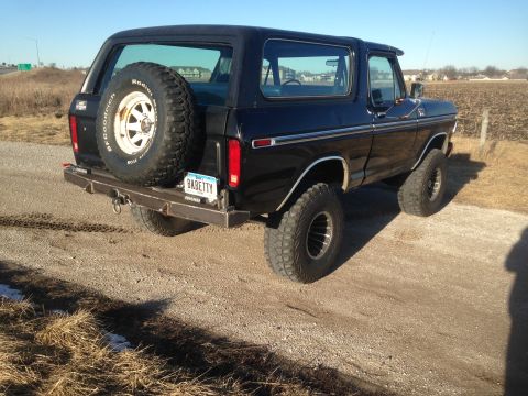 1979 Ford Bronco - The rear driver's side quarter view of a black Ford Bronco is shown parked on a gravel road. A spare tire is mounted on the rear bumper, an…