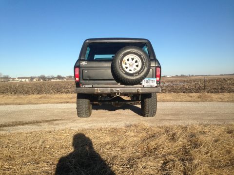 1979 Ford Bronco - The rear view of a black Ford Bronco parked on a dirt road with a spare tire mounted on the back. The vehicle has a custom bumper and a lic…