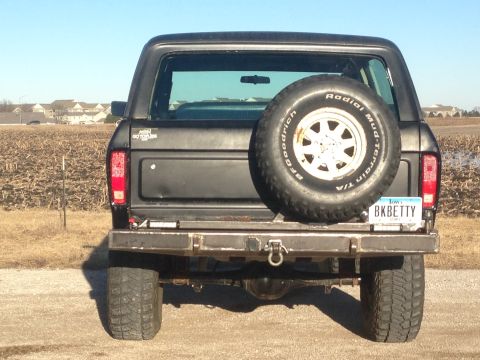 1979 Ford Bronco - The rear view of a dark colored Ford Bronco is shown, featuring a spare tire mounted on the tailgate and an Iowa license plate reading "BKB…