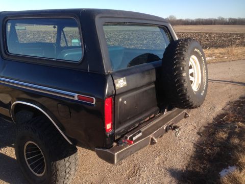 1979 Ford Bronco - The rear passenger side of a black Ford Bronco is shown parked on a dirt road with a field in the background. A spare tire is mounted on th…