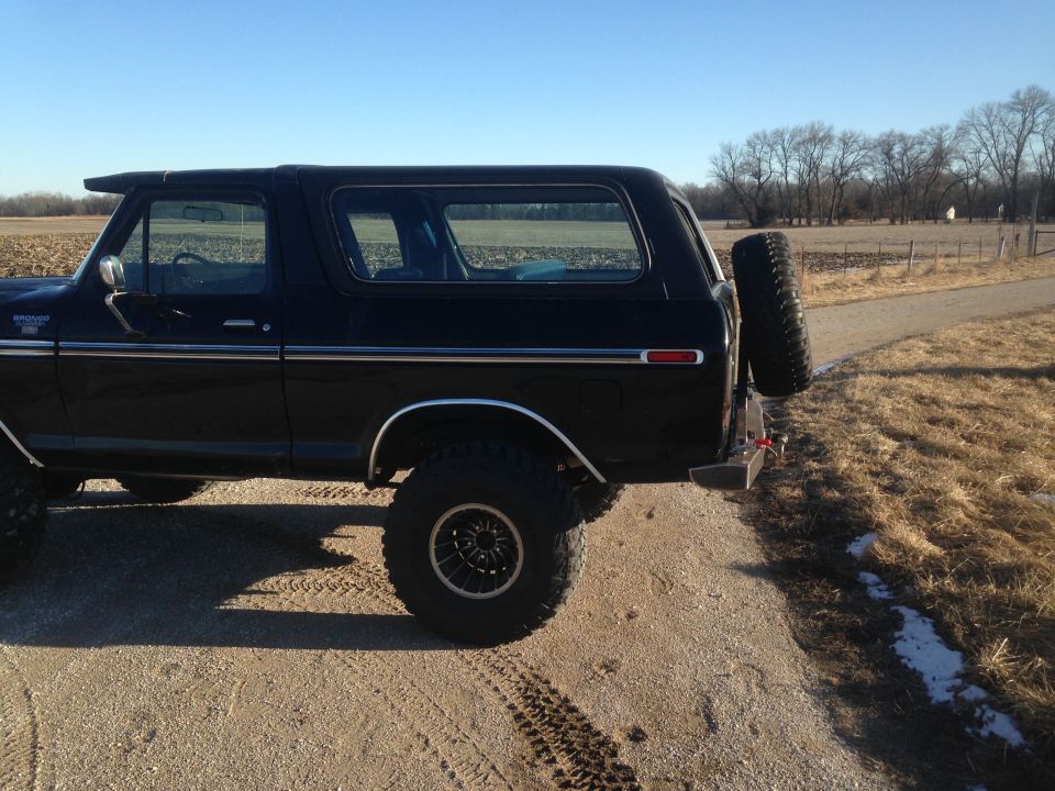 Black 1979 Ford Bronco rear driver's side view with spare tire on back, parked on dirt road.