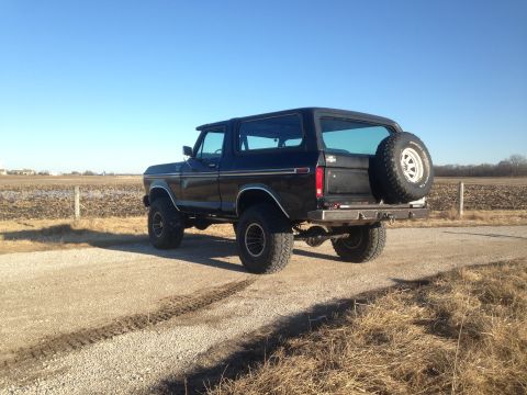 1979 Ford Bronco - The rear driver's side of a black Ford Bronco is shown parked on a gravel road with a field and fence in the background. A spare tire is mo…