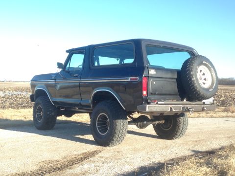 1979 Ford Bronco - The rear passenger side of a lifted black 1979 Ford Bronco is shown parked on a dirt road with a field in the background under a clear blue…