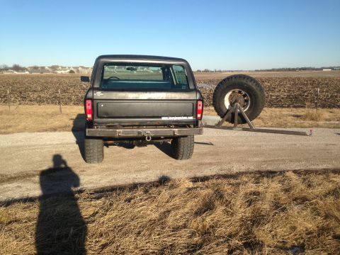 1979 Ford Bronco - Rear view of a black 1979 Ford Bronco with a custom spare tire carrier mounted to the rear bumper. The vehicle is parked on a gravel road i…